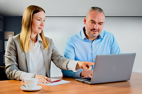 Eine junge Frau und ein Mann in Geschäftskleidung arbeiten gemeinsam an einem Laptop und lächeln.